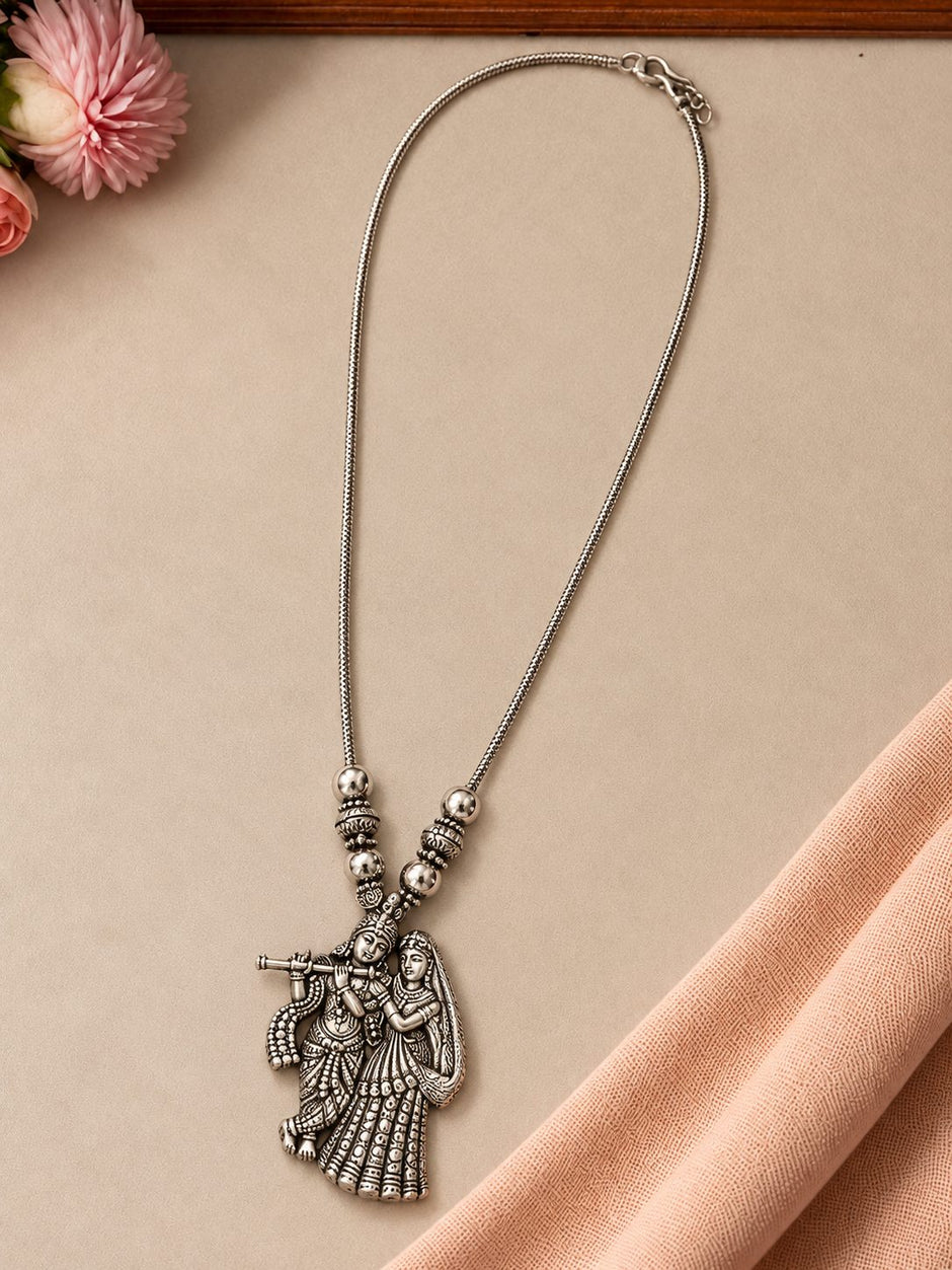 Oxidised silver Radha Krishna pendant necklace laid flat on a beige surface with pink flowers