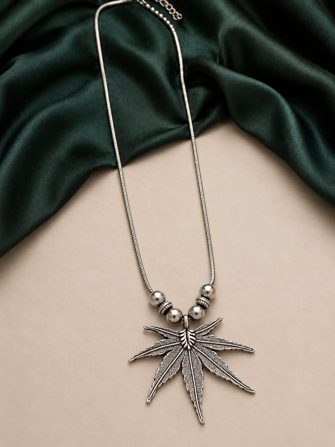 Oxidised silver leaf pendant long necklace laid flat on beige surface with green silk fabric background