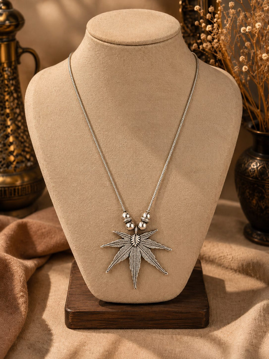 Oxidised silver leaf pendant necklace displayed on a beige jewellery bust with decorative brass props in background