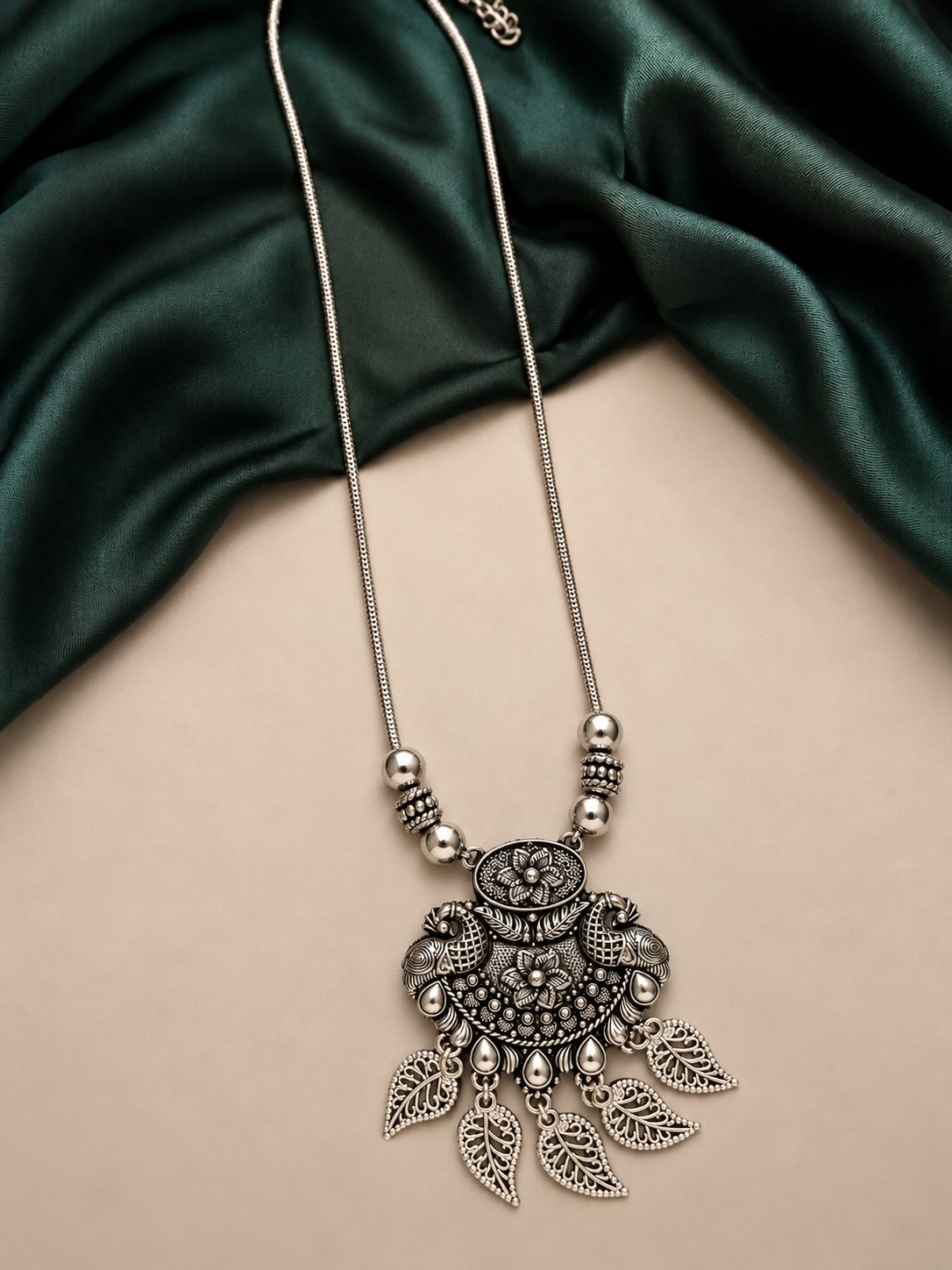 Oxidised silver pendant necklace with filigree leaf drops laid flat on green satin fabric