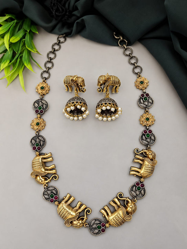 Dual-tone elephant motif necklace and jhumka earring set laid flat on a grey surface with green leaves