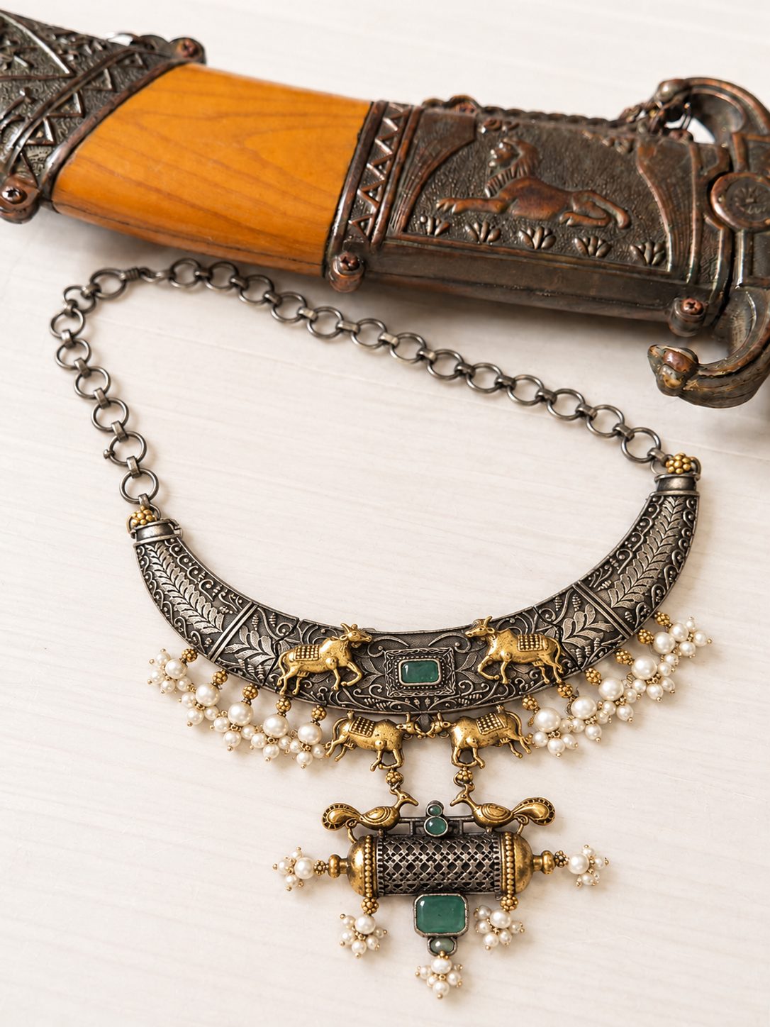 Oxidised dual-tone choker necklace with gold Nandi figurines and pearl clusters laid next to a decorative antique dagger