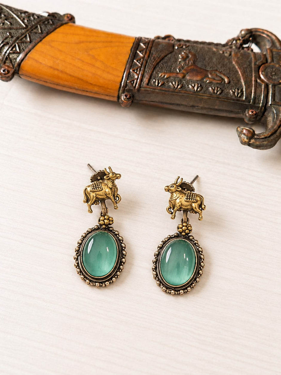 Close-up of gold-tone Nandi bull drop earrings with oval green cabochon stones on a light wooden surface