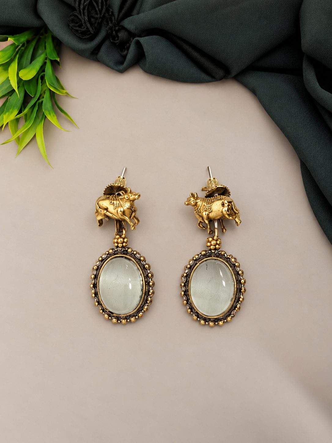 Close-up of golden cow figurine drop earrings with oval green cabochon stones on beige surface