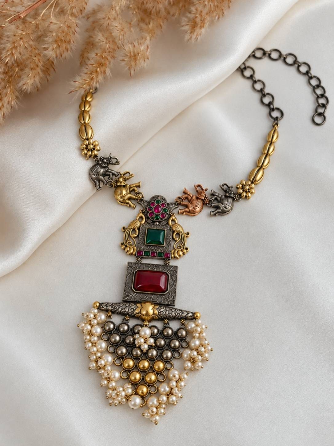 Flat-lay of oxidised dual-tone elephant necklace with ruby and emerald stone pendant and pearl cluster drop on white fabric