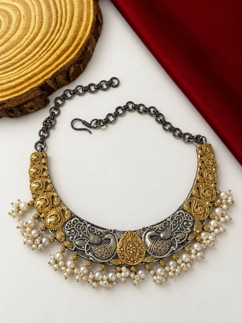 Crescent-shaped peacock choker necklace with antique gold motifs and white pearl drops laid flat on white surface