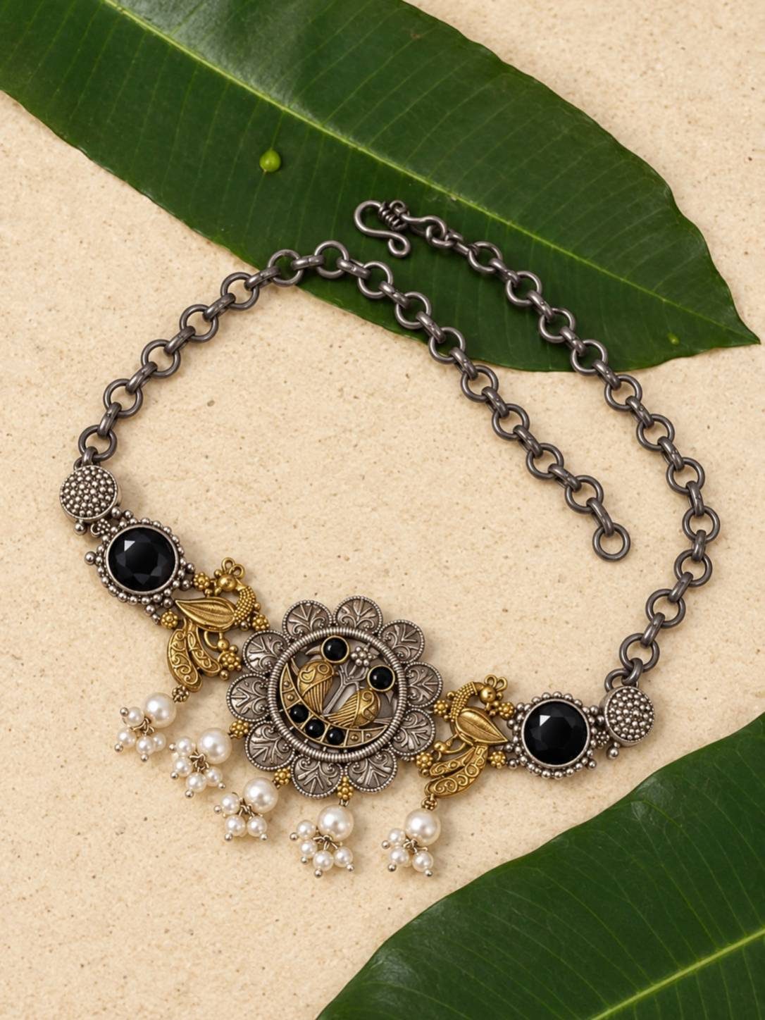 Oxidised peacock choker necklace with pearl drops laid flat on mango leaves and sandy surface