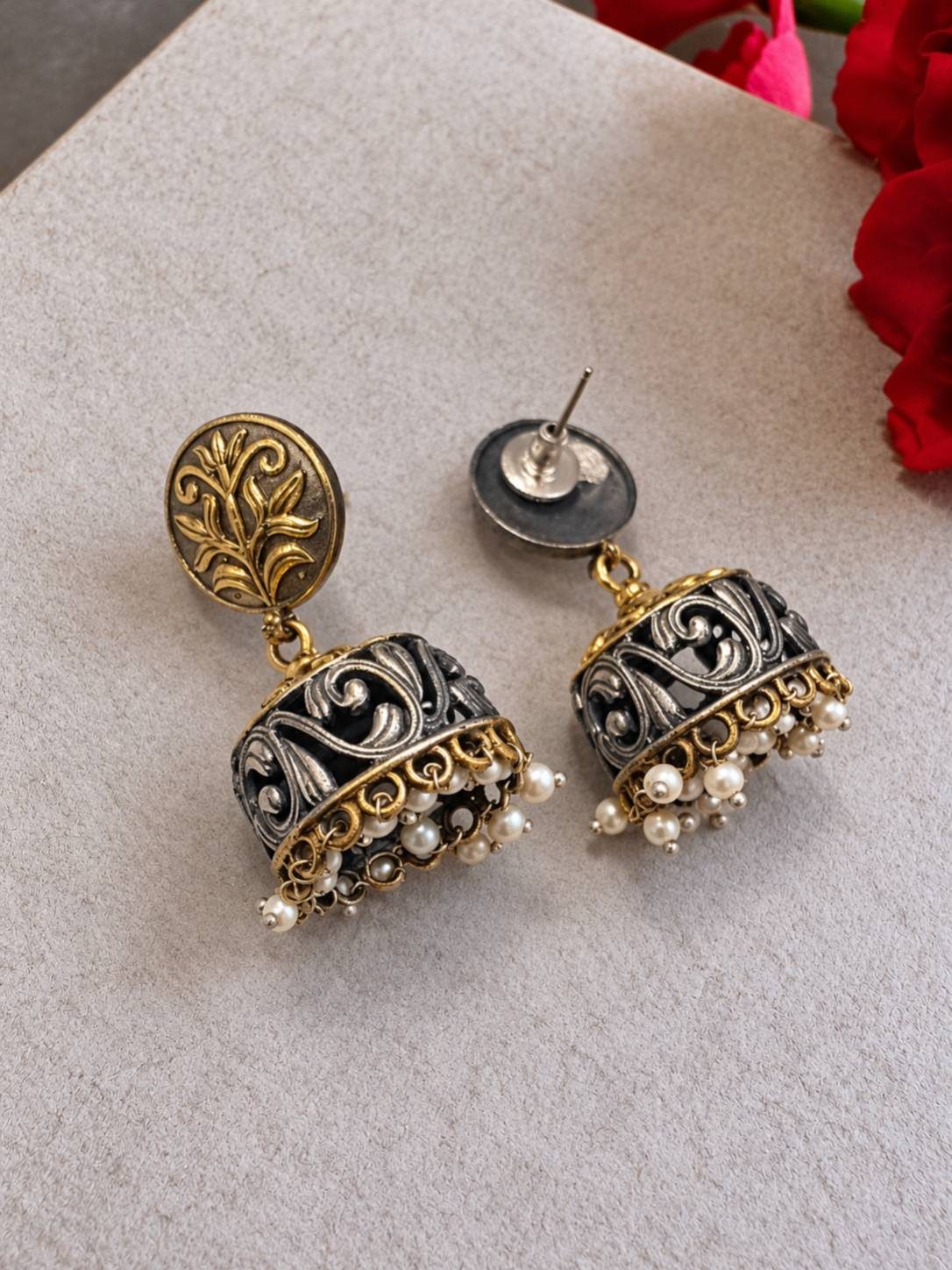 Close-up of dual-tone jhumka earrings with botanical coin stud and white pearl drops on grey surface