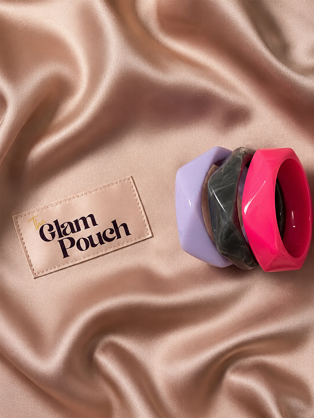 Three stacked resin bangles in marbled grey, fuchsia pink, and lavender on a satin rose gold background next to The Glam Pouch label