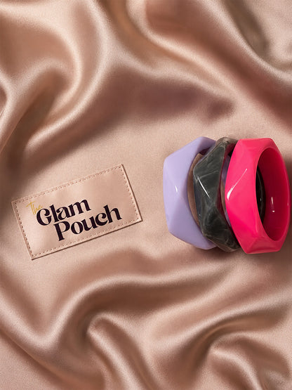 Three stacked resin bangles in marbled grey, fuchsia pink, and lavender on a satin rose gold background next to The Glam Pouch label