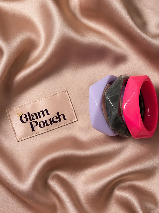 Three stacked resin bangles in marbled grey, fuchsia pink, and lavender on a satin rose gold background next to The Glam Pouch label