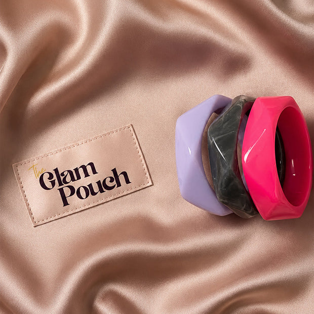 Three stacked resin bangles in marbled grey, fuchsia pink, and lavender on a satin rose gold background next to The Glam Pouch label
