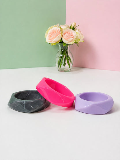 Three geometric resin bangles laid flat on a white surface in front of a pastel pink and green backdrop with roses