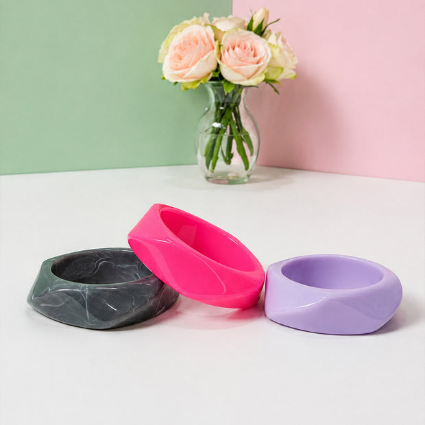 Three geometric resin bangles laid flat on a white surface in front of a pastel pink and green backdrop with roses