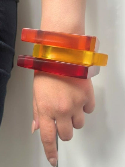 Three translucent resin square bangles in red, orange, and yellow stacked on a woman's wrist against a grey background