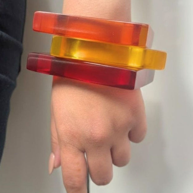 Three translucent resin square bangles in red, orange, and yellow stacked on a woman's wrist against a grey background