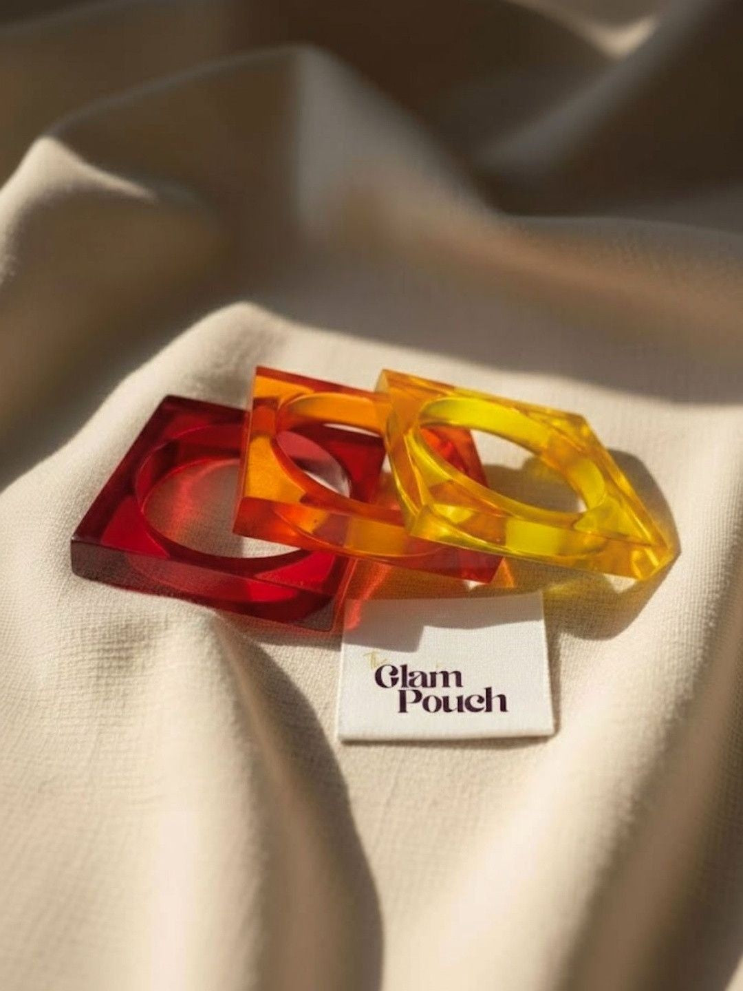 Red, orange, and yellow acrylic square bangles resting on cream fabric beside a Glam Pouch branded tag in warm sunlight