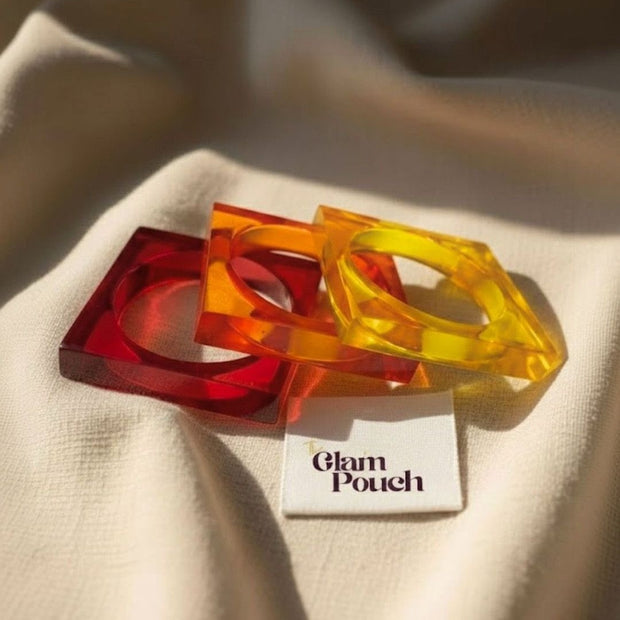 Red, orange, and yellow acrylic square bangles resting on cream fabric beside a Glam Pouch branded tag in warm sunlight