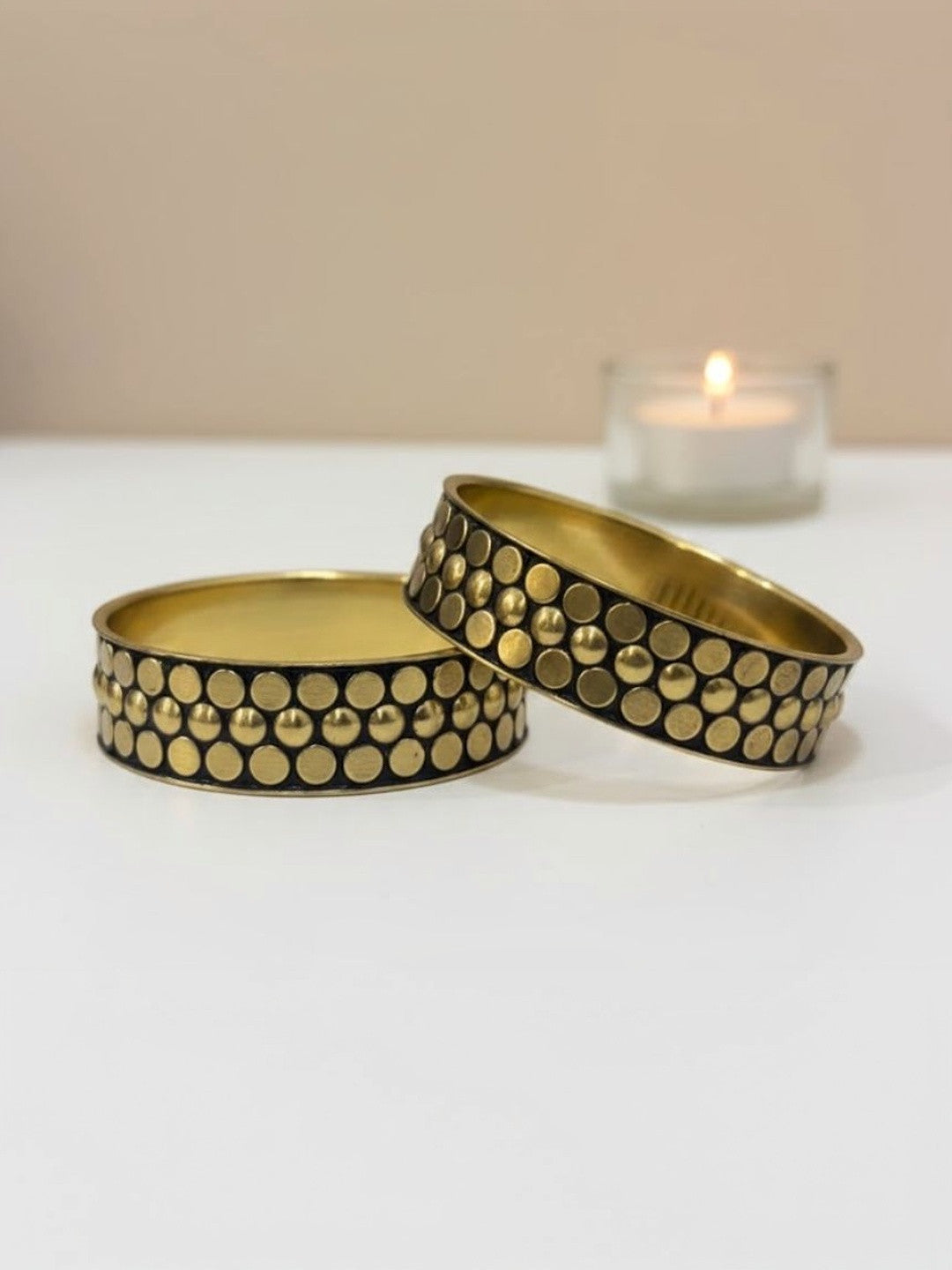 Two wide oxidised gold brass bangles with raised circular studs on a dark background, displayed flat beside a lit tealight candle