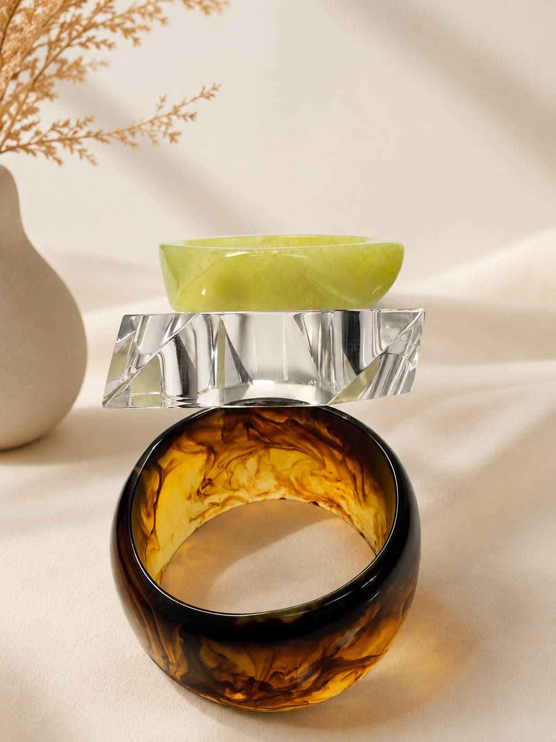 Lime green, crystal-cut clear, and tortoiseshell marbled resin bangles stacked on ivory fabric near dried botanicals