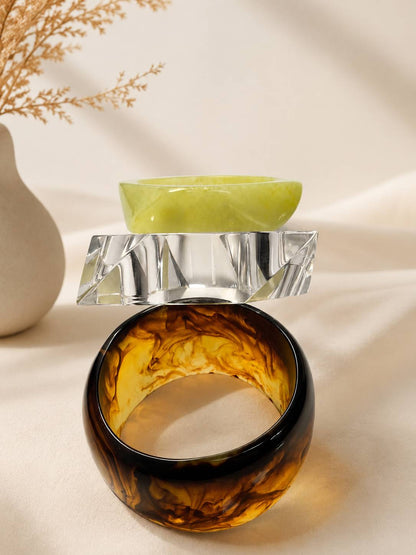 Lime green, crystal-cut clear, and tortoiseshell marbled resin bangles stacked on ivory fabric near dried botanicals