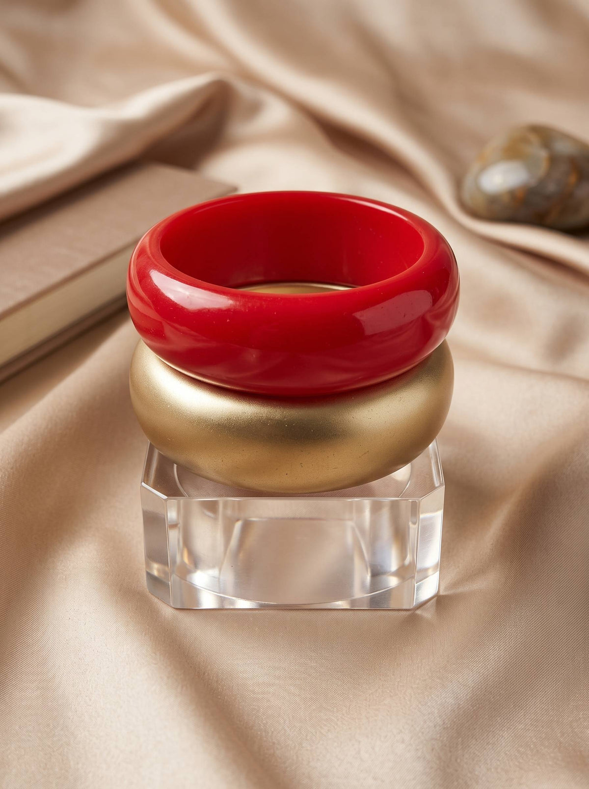 Three stacked resin bangles — red, gold and clear acrylic — displayed on a faceted crystal stand against champagne satin