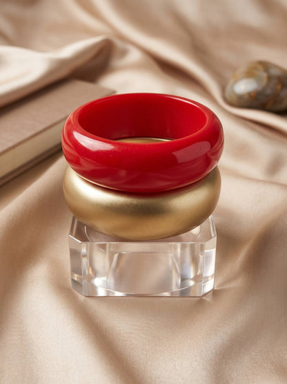 Three stacked resin bangles — red, gold and clear acrylic — displayed on a faceted crystal stand against champagne satin