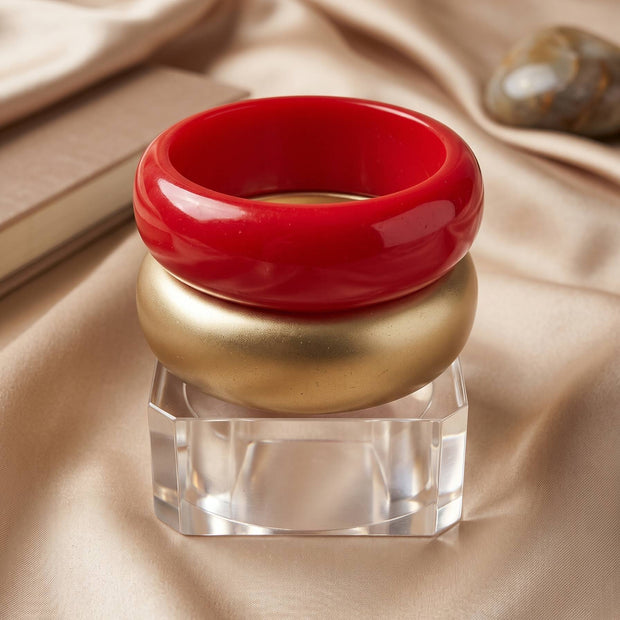 Three stacked resin bangles — red, gold and clear acrylic — displayed on a faceted crystal stand against champagne satin