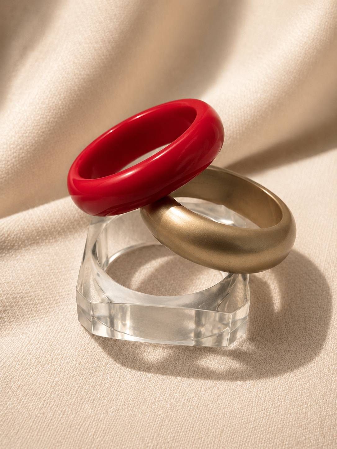 Red glossy resin bangle resting atop matte gold bangle on clear acrylic stand, angled side view on ivory fabric