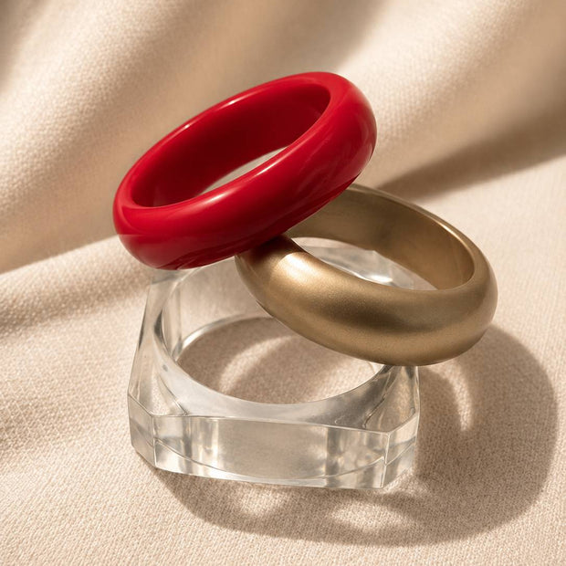 Red glossy resin bangle resting atop matte gold bangle on clear acrylic stand, angled side view on ivory fabric