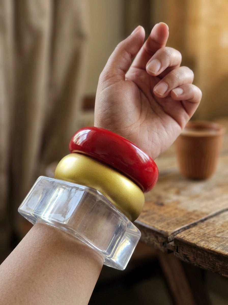 Model wearing red, gold and clear acrylic resin bangle stack on raised wrist near a rustic wooden table