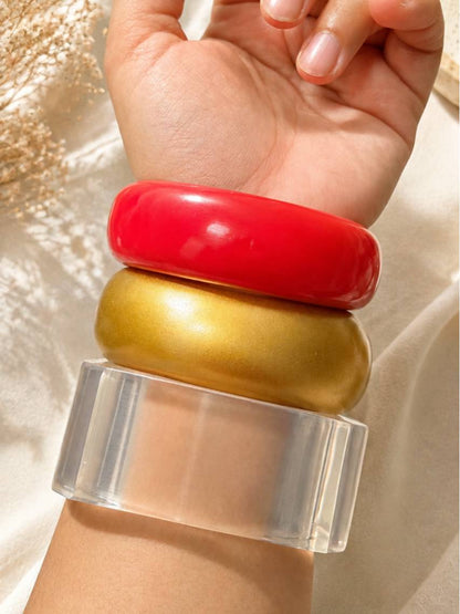 Close-up of resin bangle trio — cherry red, satin gold and transparent cuff — stacked on a model's wrist in sunlight