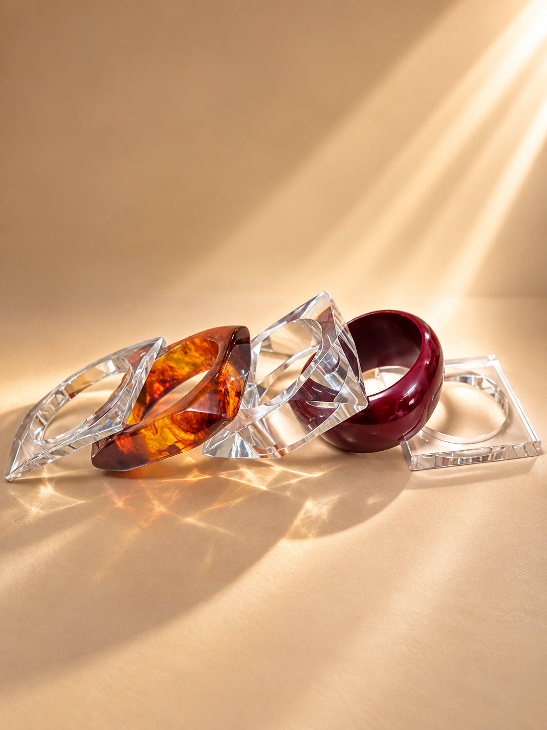 Four acrylic and resin bangles laid flat on a warm beige surface with sunlight casting prismatic shadows