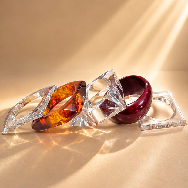 Four acrylic and resin bangles laid flat on a warm beige surface with sunlight casting prismatic shadows
