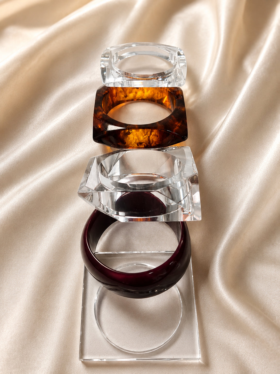 Four bangles stacked vertically on cream satin showing clear acrylic, amber tortoiseshell and burgundy resin styles