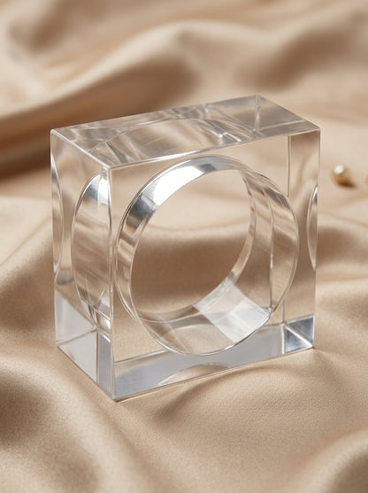 Clear acrylic square bangle with circular inner band resting on cream satin fabric