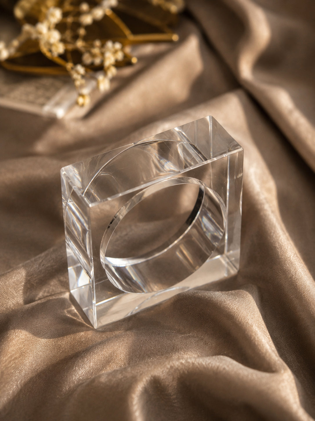 Close-up of clear acrylic square bangle on golden satin fabric with floral decor in background