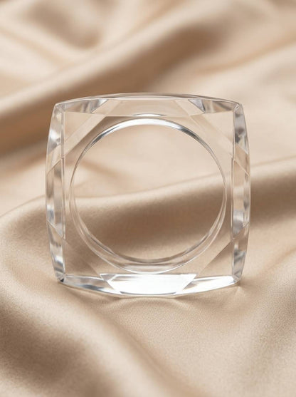 Clear acrylic square bangle laid flat on cream satin fabric, top-down view showing circular inner channel and bevelled corners