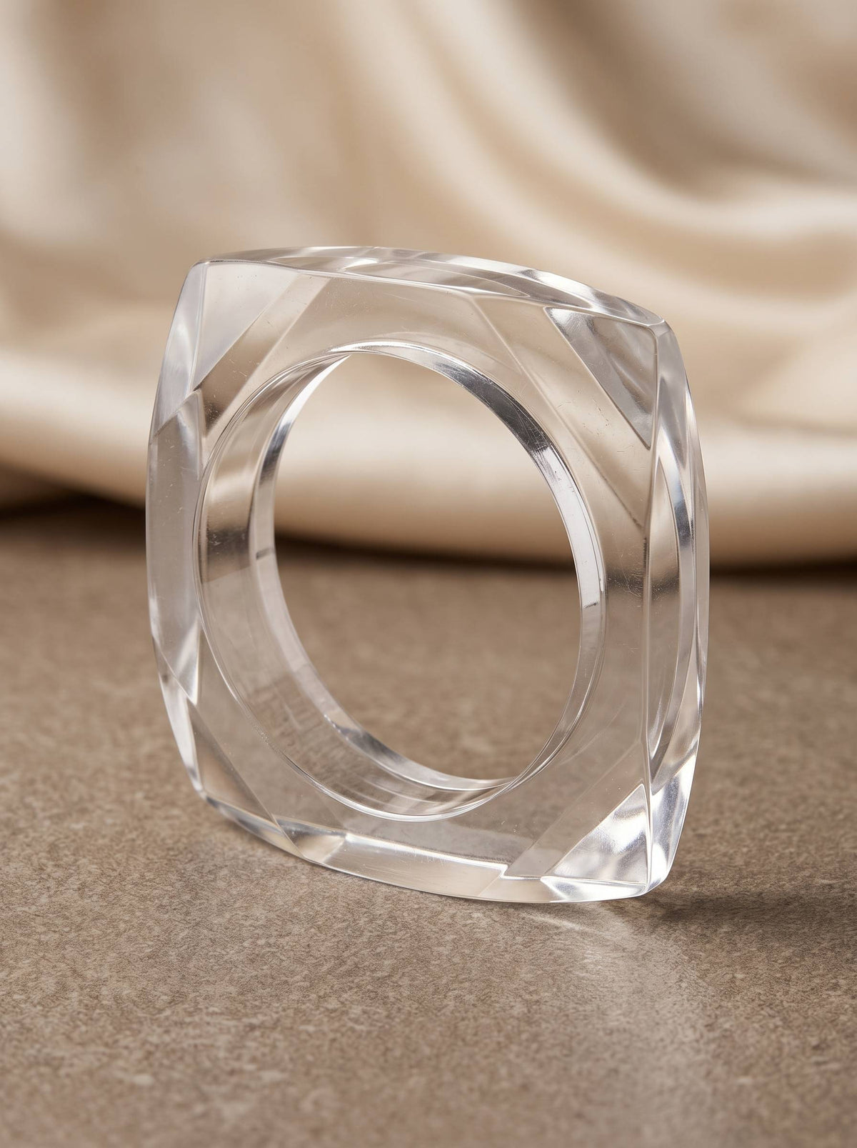 Transparent square acrylic bangle standing upright on a stone surface with cream satin draped in background showing full 3D profile