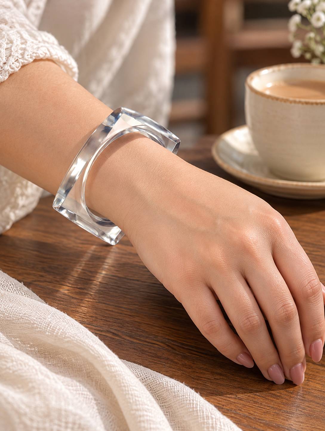 Woman wearing a clear acrylic square bangle on her wrist resting on a wooden table beside a cup of coffee