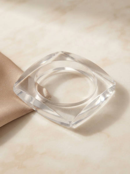 Clear faceted acrylic square bangle lying flat on a marble surface leaning against folded champagne satin fabric