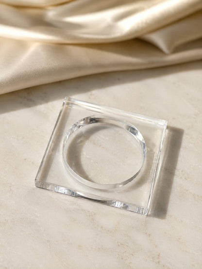 Top-down view of clear acrylic square bangle lying flat on a white marble surface with gold satin in background
