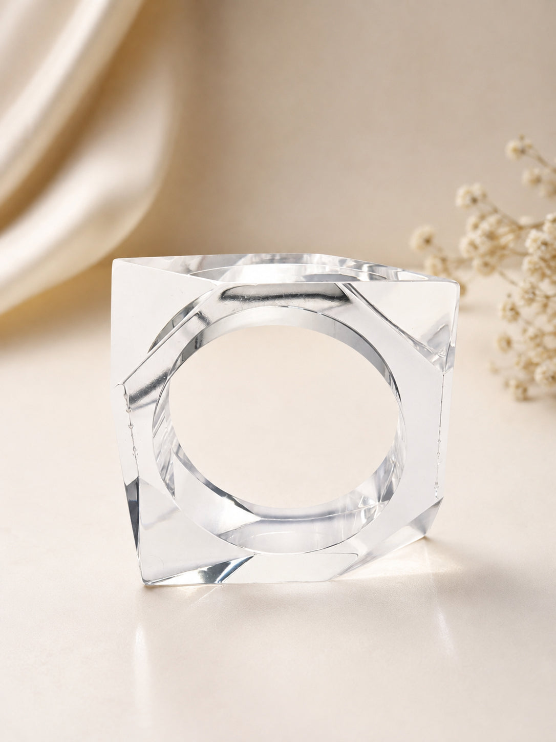 Clear acrylic square cuff bangle standing upright showing faceted corners and circular opening, shot against a cream background with dried flowers