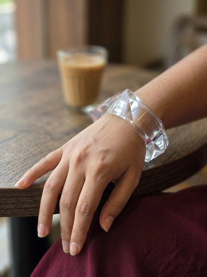 Clear acrylic square cuff bangle worn on a woman's wrist resting on a wooden table edge with a latte in the background