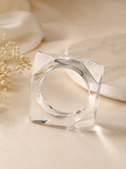 Polished clear acrylic square cuff bangle propped upright on cream satin surface beside a white stone and dried flowers