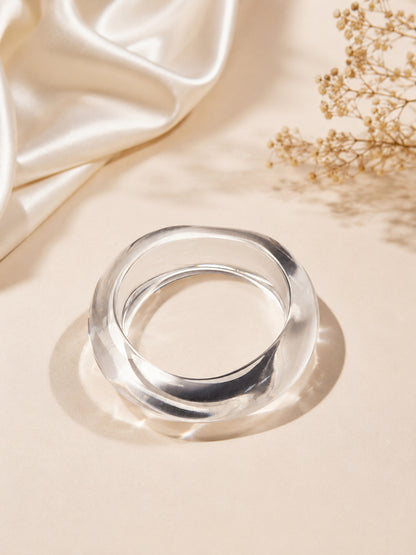 Clear acrylic bangle lying flat on a beige surface with draped satin fabric and dried baby's breath flowers nearby