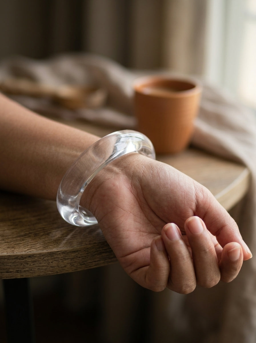 Clear acrylic bangle worn on a woman's wrist resting on the edge of a wooden table with a terracotta cup in the background