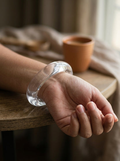 Clear acrylic bangle worn on a woman's wrist resting on the edge of a wooden table with a terracotta cup in the background