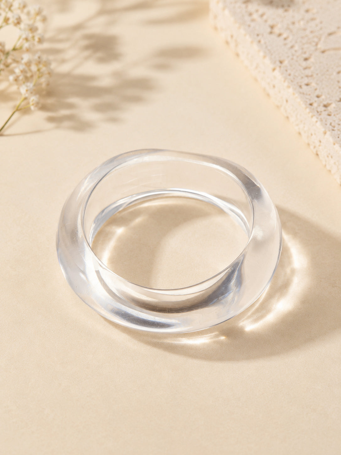 Clear acrylic twisted-silhouette bangle on a beige surface with soft shadows and dried florals in the corner