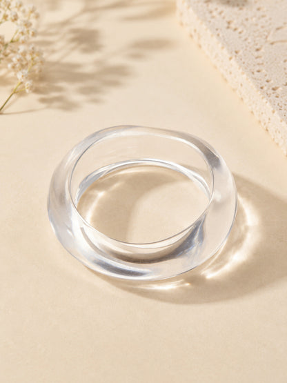 Clear acrylic twisted-silhouette bangle on a beige surface with soft shadows and dried florals in the corner